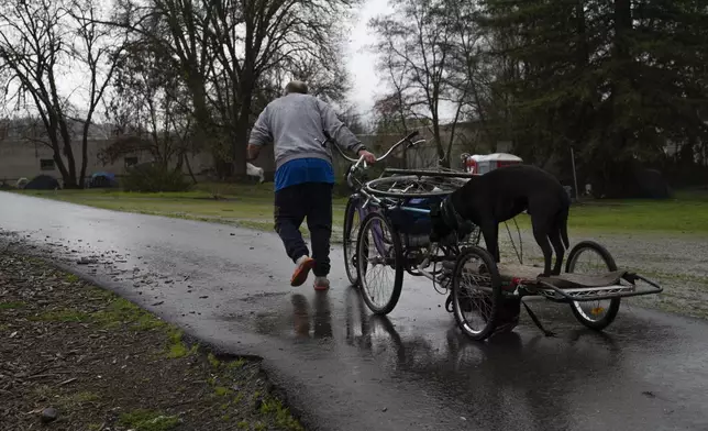 FILE - With an American flag flying in the background and tents in the distance, amputee Donnie Snow, who is homeless, uses his bike to carry his dog Bubba up a hill in the rain at Riverside Park, March 23, 2024, in Grants Pass, Ore. On Friday, June 28, the Supreme Court ruled that cities can enforce bans on homeless people sleeping outdoors in West Coast areas where shelter space is lacking. (AP Photo/Jenny Kane, File)