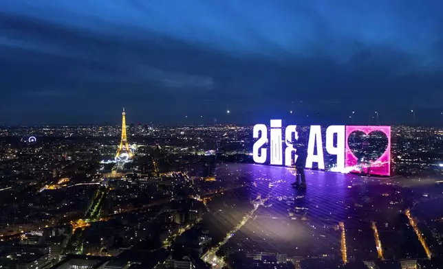 A visitor strolls past a "Paris" sign that is reflected in the glass wall of the Montparnasse tower observation deck as the Eiffel Tower stands in the background ahead of the 2024 Summer Olympics, Tuesday, July 23, 2024, in Paris. (AP Photo/David Goldman)