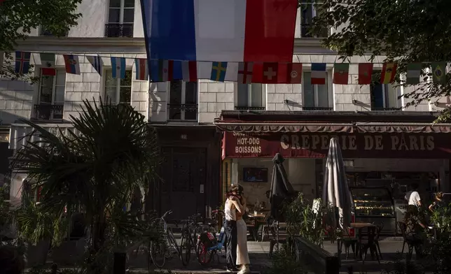 A couple embraces in the street as the flag of France hangs from a restaurant ahead of the 2024 Summer Olympics, Sunday, July 21, 2024, in Paris. (AP Photo/David Goldman)