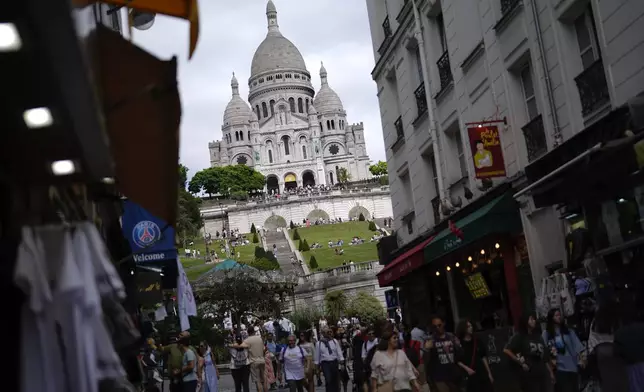 People walk past souvenir shops and restaurants below Sacre Coeur of Montmartre Basilica, ahead of the 2024 Summer Olympics, Monday, July 22, 2024, in Paris, France. (AP Photo/Rebecca Blackwell)