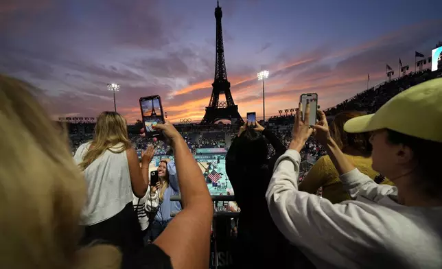 Spectators photograph a colorful sunset at Eiffel Tower Stadium prior to a beach volleyball match between The United States and Canada at the 2024 Summer Olympics, Saturday, July 27, 2024, in Paris, France. (AP Photo/Robert F. Bukaty)