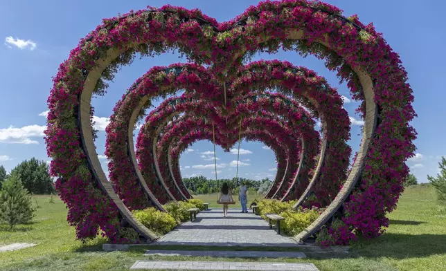 A woman swings from a heart-shaped flower installation in Dobro Park, Motyzhyn, Kyiv region, Ukraine, Wednesday, June 26, 2024. Despite hardships brought by war flowers fill Kyiv and other Ukrainian cities. They burst out of planters that line the capital's backroads and grand boulevards and are fixed to lampposts. (AP Photo/Anton Shtuka)