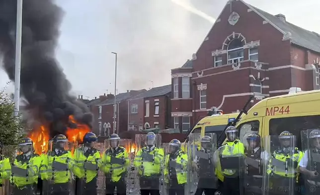 A police van, rear, burns as police line up after clashing with an unruly crowd, Tuesday, July 30, 2024, in Southport, northwest England, near where three girls were stabbed to death in a dance class the day before. The violence erupted shortly after a peaceful vigil was attended by hundreds in the center of Southport to mourn the 13 victims of the stabbings, including seven still in critical condition. (Pat Hurst/PA via AP)