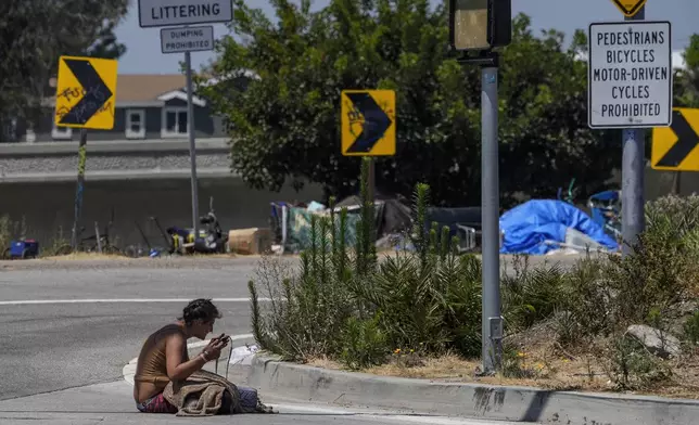 A homeless woman sits on the street at the entrance to an off-ramp to the CA-105 freeway next to a homeless encampment Friday, July 26, 2024, in Los Angeles. (AP Photo/Damian Dovarganes)