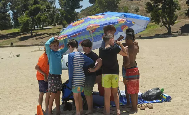 A group of youths shelter in the shade after being asked by lifeguards to retrieve their footwear before getting onto the sunny beach area to prevent burnt feet at Castaic Lake beach as the temperature rises Monday, July 8, 2024, in Castaic, Calif. (AP Photo/Damian Dovarganes)