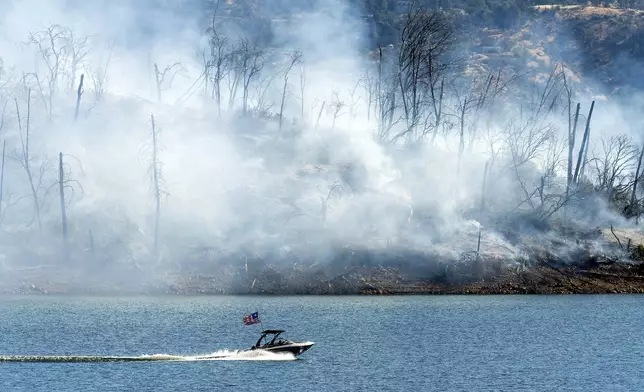 A boat crosses Lake Oroville with a smoldering hillside behind as the Thompson Fire burns in Oroville, Calif., on Wednesday, July 3, 2024. An extended heatwave blanketing Northern California has resulted in red flag fire warnings and power shutoffs. (AP Photo/Noah Berger)