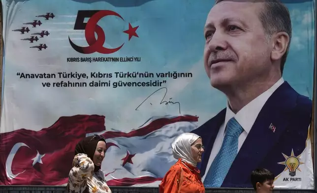 Turkish Women and a kid walk in front of a poster of President Recep Tayyip Erdogan after a military parade, in the Turkish occupied area of the divided capital Nicosia, Cyprus, Saturday, July 20, 2024. (AP Photo/Petros Karadjias)