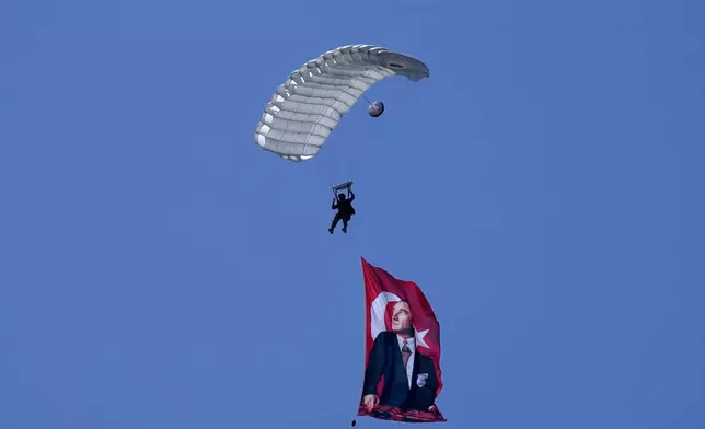 A Turkish parachuter prepares for a landing as fly over the military parade with a Turkish flag and portrait of the Kemal Ataturk, the founder of modern Turkey, during a military parade, in the Turkish occupied area of the divided capital Nicosia, Cyprus, Saturday, July 20, 2024. The tinny howl of air raid sirens echoed across ethnically divided Cyprus at the crack of dawn on Saturday, marking 50 years from the day that changed the island nation's history. It's heard every year as the Greek Cypriot controlled south laments the date Turkish troops invaded in the wake of a coup by supporters of union with Greece. (AP Photo/Petros Karadjias)