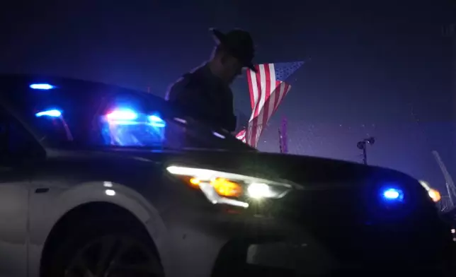 An officer stands at his car early Sunday, July 14, 2024, closing access to the site of the rally where former President Donald Trump was the target of an assassination attempt on Saturday in Butler, Pa.. Behind the officer, a large flag from the rally still flies. (AP Photo/Sue Ogrocki)