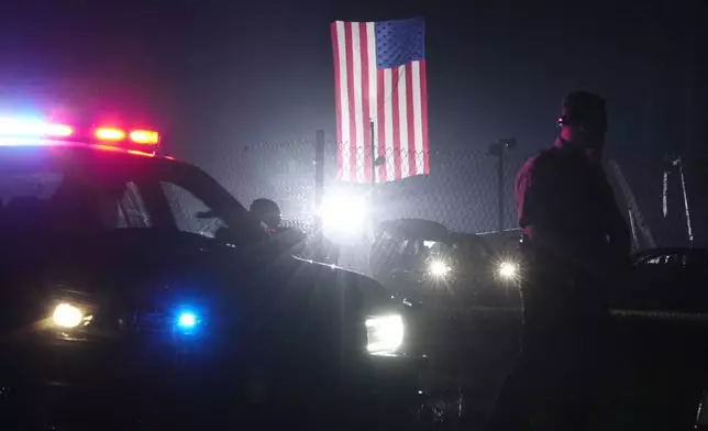 An officer stands at his car early Sunday, July 14, 2024, closing access to the site of the rally where former President Donald Trump was the target of an assassination attempt on Saturday in Butler, Pa.. Behind the officer, a large flag from the rally still flies. (AP Photo/Sue Ogrocki)