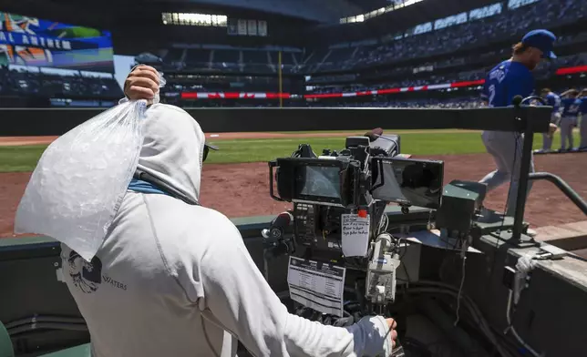 A camera operator, left, uses a bag of ice to stay cool as Toronto Blue Jays designated hitter Justin Turner, right, enters the dugout as temperatures rise before a baseball game against the Seattle Mariners, Sunday, July 7, 2024, in Seattle. (AP Photo/Jason Redmond)