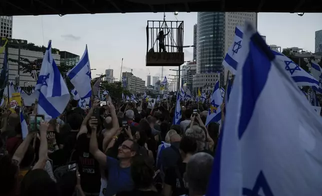 A demonstrator stands in a cage hanging from a pedestrian bridge as others wave Israeli flags during a protest marking nine months since the start of the war and calling for the release of hostages held in the Gaza Strip by the Hamas militant group, in Tel Aviv, Israel, Sunday, July 7, 2024. (AP Photo/Leo Correa)