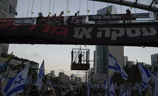 A demonstrator stands in a cage hanging from a pedestrian bridge as others wave Israeli flags during a protest marking nine months since the start of the war and calling for the release of hostages held in the Gaza Strip by the Hamas militant group, in Tel Aviv, Israel, Sunday, July 7, 2024. (AP Photo/Leo Correa)