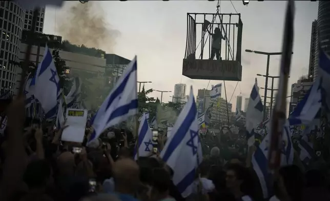 A demonstrator stands in a cage hanging from a pedestrian bridge as others wave Israeli flags during a protest marking nine months since the start of the war and calling for the release of hostages held in the Gaza Strip by the Hamas militant group, in Tel Aviv, Israel, Sunday, July 7, 2024. (AP Photo/Leo Correa)