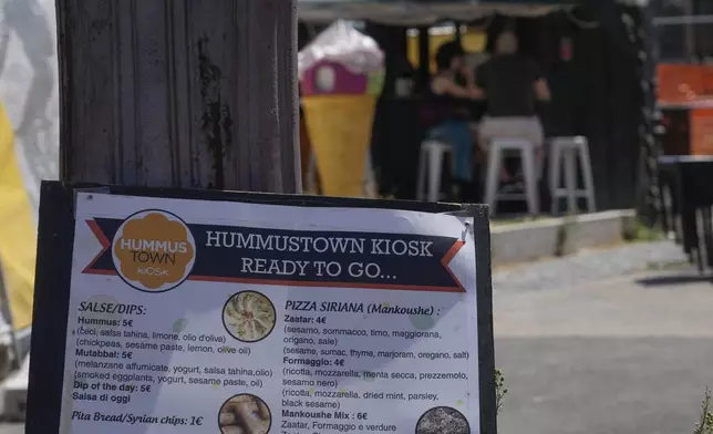 A menu is displayed outside a HummusTown kiosk in Rome, Saturday, July 27, 2024. A pair of Syrians have created community that provides support to migrants and vulnerable people in Rome, by sharing the flavors of a homeland torn by civil war. Created in 2018 as a "humanitarian catering service," HummusTown originally aimed at raising funds for families and friends in Syria. (AP Photo/Gregorio Borgia)