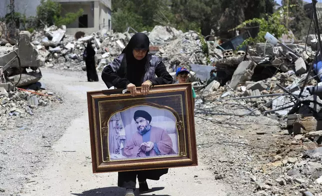 A woman carries a frame that shows a portrait for Hezbollah leader Sayyid Hassan Nasrallah, as she passes destroyed houses that were hit by Israeli airstrikes, in Aita al-Shaab, a Lebanese border village with Israel, south Lebanon, Saturday,, June 29, 2024. (AP Photo/Mohammed Zaatari)