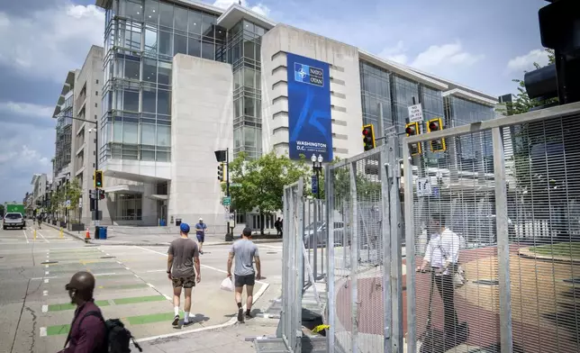 People walk and ride scooters through security fencing, by the Walter E. Washington Convention Center, which has been draped with signage ahead of the NATO Summit, Monday, July 8, 2024. (AP Photo/Jacquelyn Martin)