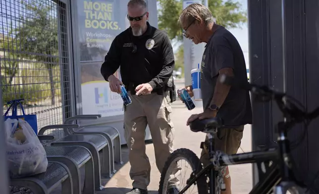 Mark Paulson, a Public Response and Code Enforcement officer, gives cans of cold water to Russell Eibeck, Wednesday, July 10, 2024, in Henderson, Nev. About 14 officers from the Office of Public Response drove around the city Wednesday, offering water, electrolytes, free bus tickets, and rides to cooling centers during a heat emergency. (AP Photo/John Locher)