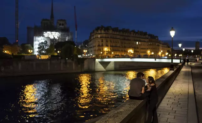 Victor, left, and his girlfriend, Florence, talk along the Seine River at dusk as the spire of the Notre Dame Cathedral rises in the background ahead of the 2024 Summer Olympics, Tuesday, July 16, 2024, in Paris, France. (AP Photo/David Goldman)