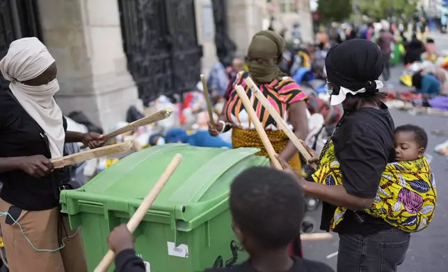 Ivory Coast's Achata Traore, right, with her daughter Fatima on her back, drums on bins during a protest at the 2024 Summer Olympics, Thursday, July 25, 2024, in Paris, France. On the eve of the grandiose opening ceremony for the Paris Olympics, police cleared out migrants sleeping in a tent camp in the capital as social and environmental advocacy groups raise attention to criticisms of the Paris Games such as the displacement of migrants and housing issues. (AP Photo/David Goldman)