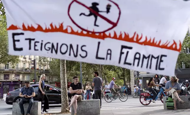 Pedestrians and bicyclists pass a sign reading "Let's extinguish the flame," hanging in Republique plaza during a demonstration by several associations for what they called the "Counter Opening Ceremony" against the 2024 Summer Olympics, Thursday, July 25, 2024, in Paris. Demonstrators gave speeches about the cost of the Olympic Games and said authorities have been using the Games as a pretext for what they call social cleansing, by removing migrants and homeless people off the streets in order to preserve a picture-postcard image of the city for the millions of visitors coming. (AP Photo/David Goldman)