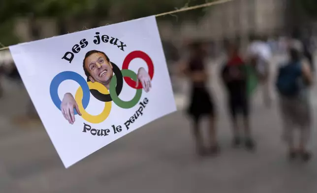 An image of French President Emmanuel Macron and the Olympic rings decorates a sign reading "The games for the people," hanging in Republique plaza during a demonstration by several associations for what they called the "Counter Opening Ceremony" against the 2024 Summer Olympics, Thursday, July 25, 2024, in Paris. Demonstrators gave speeches about the cost of the Olympic Games and said authorities have been using the Games as a pretext for what they call social cleansing, by removing migrants and homeless people off the streets in order to preserve a picture-postcard image of the city for the millions of visitors coming. (AP Photo/David Goldman)