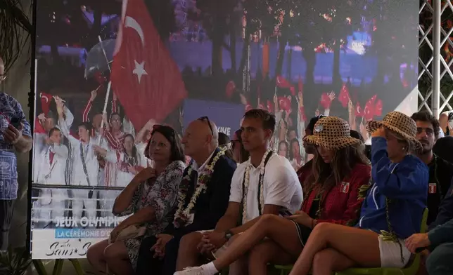 Surfers and dignitaries look on as images from the opening ceremony in Paris are shown on a screen during an opening ceremony for the 2024 Summer Olympics surfing competition Friday, July 26, 2024, in Papara, Tahiti. (AP Photo/Gregory Bull)