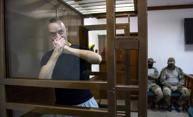 FILE - Ivan Safronov, an adviser to the director of Russia's state space agency, greets journalists while standing in a defendant’s cage in a courtroom in Moscow, Russia, Tuesday, Sept. 15, 2020, with two Federal Security Service officers sitting nearby. Safronov, a former military affairs journalist, was convicted of treason and sentenced to 22 years in prison. Authorities accused him of passing military secrets to Czech intelligence and a German national, which he denied. The case has been widely viewed as retaliation for his reporting. (AP Photo/Alexander Zemlianichenko, File)