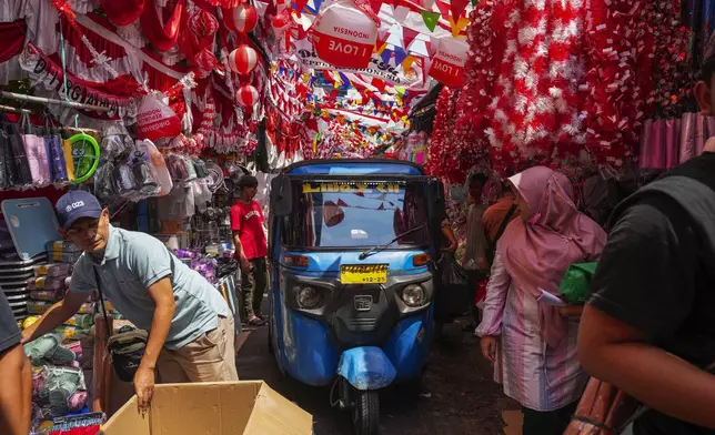 A three-wheeled motorized vehicle drives past people in an alley where vendors have displayed national flags and decoration material for sale ahead of country's Independence anniversary in Jakarta, Indonesia, Tuesday, Aug. 13, 2024. (AP Photo/Tatan Syuflana)