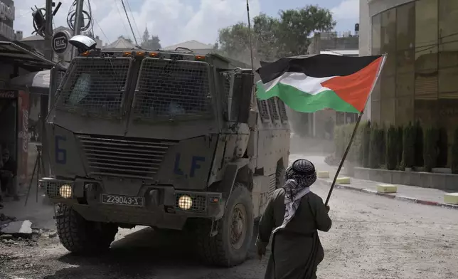 A man waves a Palestinian flag as an Israeli armored vehicle moves on a street during a military operation in the West Bank refugee camp of Nur Shams, Tulkarem, Thursday, Aug. 29, 2024. (AP Photo/Majdi Mohammed)