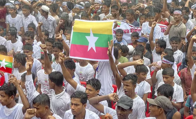 Rohingya refugees gather in the rain, with a flag of Myanmar seen at center, to demand safe return to Myanmar's Rakhine state as they mark the seventh anniversary of their mass exodus at their refugee camp at Kutupalong in Cox's Bazar district, Bangladesh, Sunday, Aug. 25, 2024. (AP Photo/ Shafiqur Rahman)