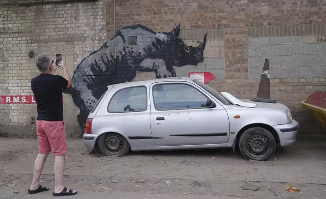 A man photographs a new artwork unveiled by Banksy, depicting a rhinoceros which looks as though it is climbing on top of a car, the eighth artwork in his animal-themed collection, on Westmoor Street in Charlton, south east London, Monday, Aug. 12, 2024. (Lucy North/PA via AP)