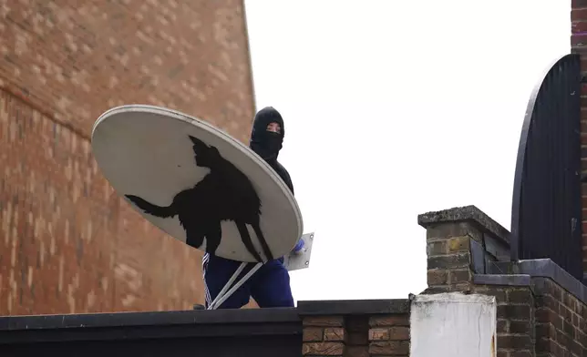 A covered-face person removes a new artwork by Banksy, depicting a howling wolf painted on a satellite dish that was placed on a shop roof in Peckham, south London, Thursday Aug. 8, 2024. (Jordan Pettitt/PA via AP)