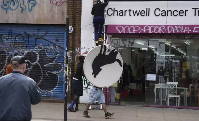 People remove a new artwork by Banksy, depicting a howling wolf painted on a satellite dish that was placed on a shop roof in Peckham, south London, Thursday Aug. 8, 2024. (Jordan Pettitt/PA via AP)