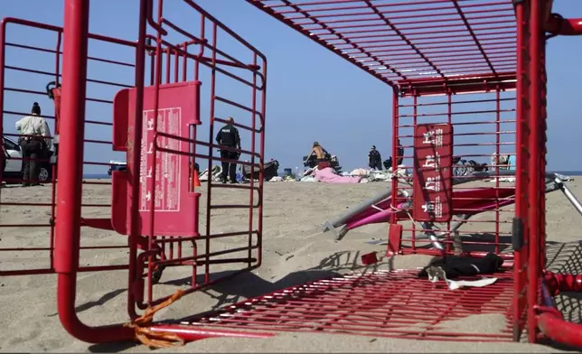 Seen through a toppled shopping cart, police and beach workers remove homeless encampments at the Dockweiler State Beach in Playa del Rey, Los Angeles, on Thursday, Aug. 22, 2024. (AP Photo/Damian Dovarganes)