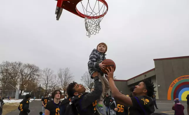 This photo provided by Spokane Public Schools shows John R. Rogers High School football players lifting a Longfellow Elementary student to the hoop during a visit to the school, Nov. 2022. Longfellow kindergarteners inspired the field trip when they wrote a collaborative letter to the older kids, saying how proud they were of the neighborhood team for earning their first win in three years, in Spokane, Wash. (Spokane Public Schools via AP)