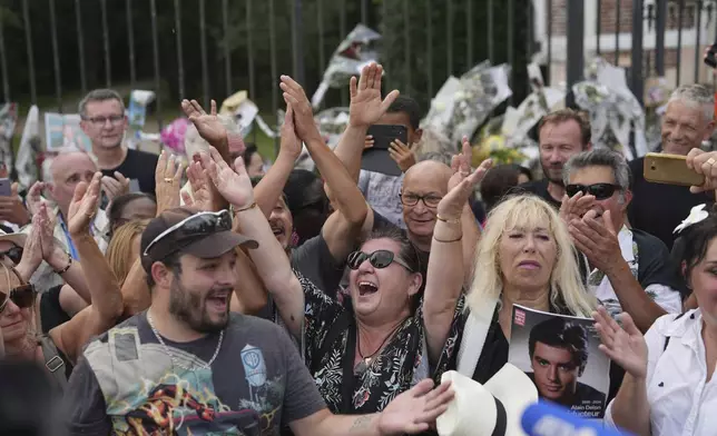 People sing a song as they pay tribute to late French actor Alain Delon, at the entrance gate of Delon's property, in Douchy, central France, Saturday, Aug. 24, 2024. (AP Photo/Thibault Camus)