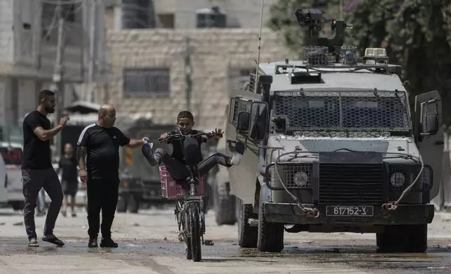 A youth rides his bicycle past an Israeli armoured vehicle during a military operation in the West Bank city of Jenin, Wednesday, Aug. 28, 2024. (AP Photo/Majdi Mohammed)