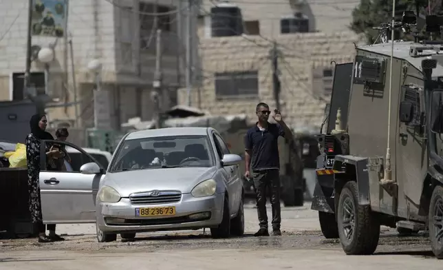 A man waves to members of the Israeli forces inside an armoured vehicle, as he stands next to his car with a woman holding a baby during a military operation in the West Bank city of Jenin, Wednesday, Aug. 28, 2024. (AP Photo/Majdi Mohammed)