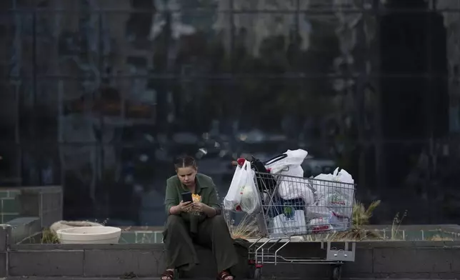 A woman looks at her phone while eating a sandwich next to a shopping cart in Haifa, Israel, Thursday, Aug. 15, 2024. Israel's economy is suffering from the nearly 11-month war with Hamas, as its leaders grind ahead with its offensive in Gaza that threatens to escalate into a wider conflict. (AP Photo/Leo Correa)