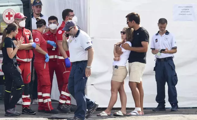 A man and a woman wait outside a tent where the body of one of the victims of a shipwreck was taken by rescuers, in Porticello, Sicily, southern Italy, Thursday, Aug. 22, 2024. Divers searching the wreck of the superyacht Bayesian that sank off Sicily on Monday recovered a fifth body on Thursday and continued to search for one more as investigators sought to learn why the vessel sank so quickly. The sign in Italian at right reads: 'Space reserved for the judicial authority and the medical examiner'. (AP Photo/Salvatore Cavalli)