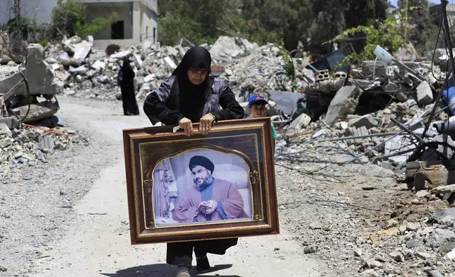 FILE - A woman carries a frame that shows a portrait for Hezbollah leader Sayyid Hassan Nasrallah, as she passes by destroyed houses that were hit by Israeli airstrikes, in Aita al-Shaab, a Lebanese border village with Israel, south Lebanon, June 29, 2024. (AP Photo/Mohammed Zaatari, File)