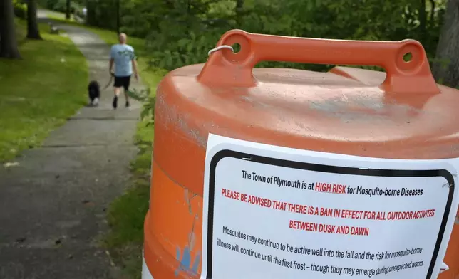 A passer-by walks a dog, Monday, Aug. 26, 2024, along a walkway, in Plymouth, Mass., near a sign that advises people of a ban in effect for outdoor activity between dusk and dawn due to the risk of exposure to mosquito-borne diseases. (AP Photo/Steven Senne)