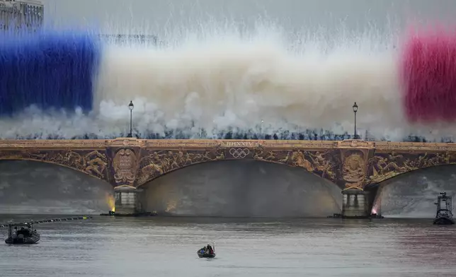 The Olympic torch travels by boat as ceremonial smoke in the colors of the France flag appear over the Seine River Paris, France, during the opening ceremony of the 2024 Summer Olympics, Friday, July 26, 2024. (AP Photo/Matthias Schrader)