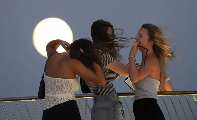 German girls on vacation in Portugal react to the strong wind while watching a nearly full supermoon rise over the Tagus river from the roof the Museum of Art, Architecture and Technology in Lisbon, Sunday, Aug. 18, 2024. (AP Photo/Armando Franca)