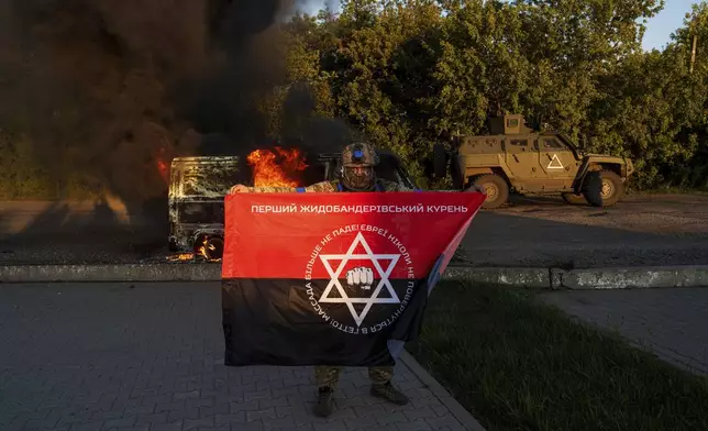 A Ukrainian serviceman holds a flag for his fallen comrade in front of a burned vehicle near Russian-Ukrainian border, Sumy region, Ukraine, Wednesday, Aug. 14, 2024. (AP Photo/Evgeniy Maloletka)