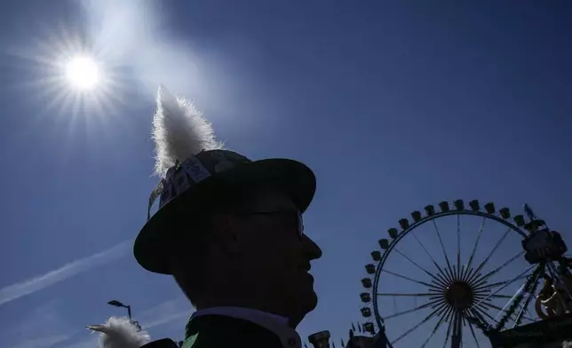 A man with a traditional Bavarian hat is pictured in the sun at start of the 189th 'Oktoberfest' beer festival in front of paintings showing Munich landmarks in Munich, Germany, Saturday, Sept. 21, 2024. (AP Photo/Matthias Schrader)