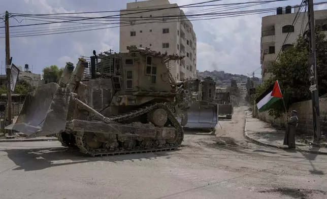 A man waves the Palestinian flag as a convoy of Israeli military bulldozers drive by during an army raid in Jenin, West Bank, Monday, Sept. 2, 2024. (AP Photo/Majdi Mohammed)