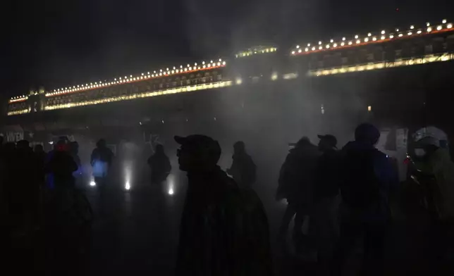 Youths are silhouetted in clouds of smoke caused by fire balls thrown at the barriers protecting the facade of the National Palace during a demonstration marking the 10-year anniversary of the disappearance of 43 students from an Ayotzinapa rural teacher's college, in Mexico City, Thursday, Sept. 26, 2024. (AP Photo/Fernando Llano)