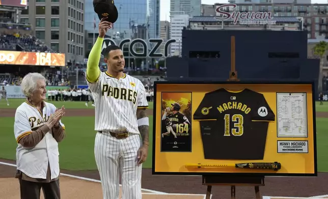 San Diego Padres' Manny Machado tips his hat to the crowd after receiving memorabilia to honor breaking the team record for all-time home runs as he stands alongside Casey Colbert, the the widow of former record holder Nate Colbert, before a baseball game against the Houston Astros Tuesday, Sept. 17, 2024, in San Diego. (AP Photo/Gregory Bull)